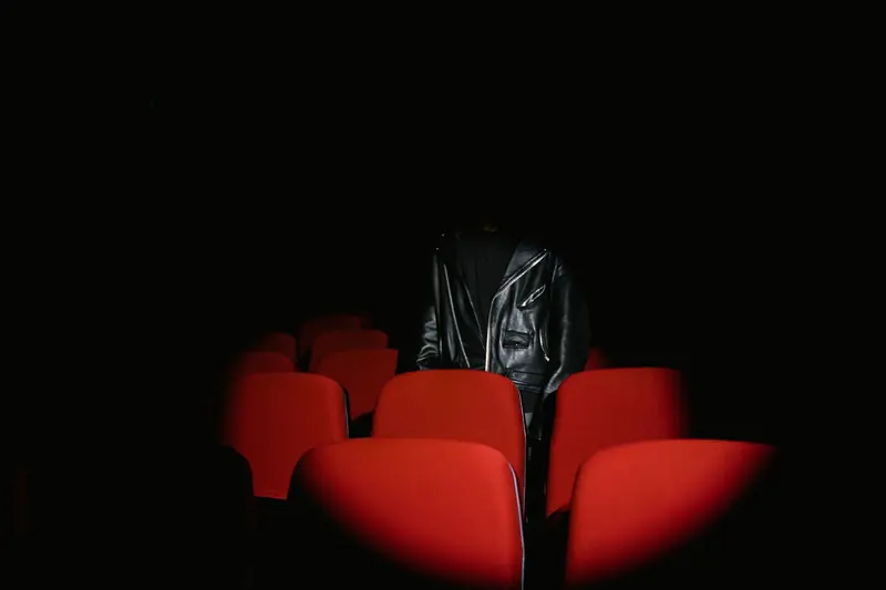 A person in a leather jacket stands among red theater seats in a dark room